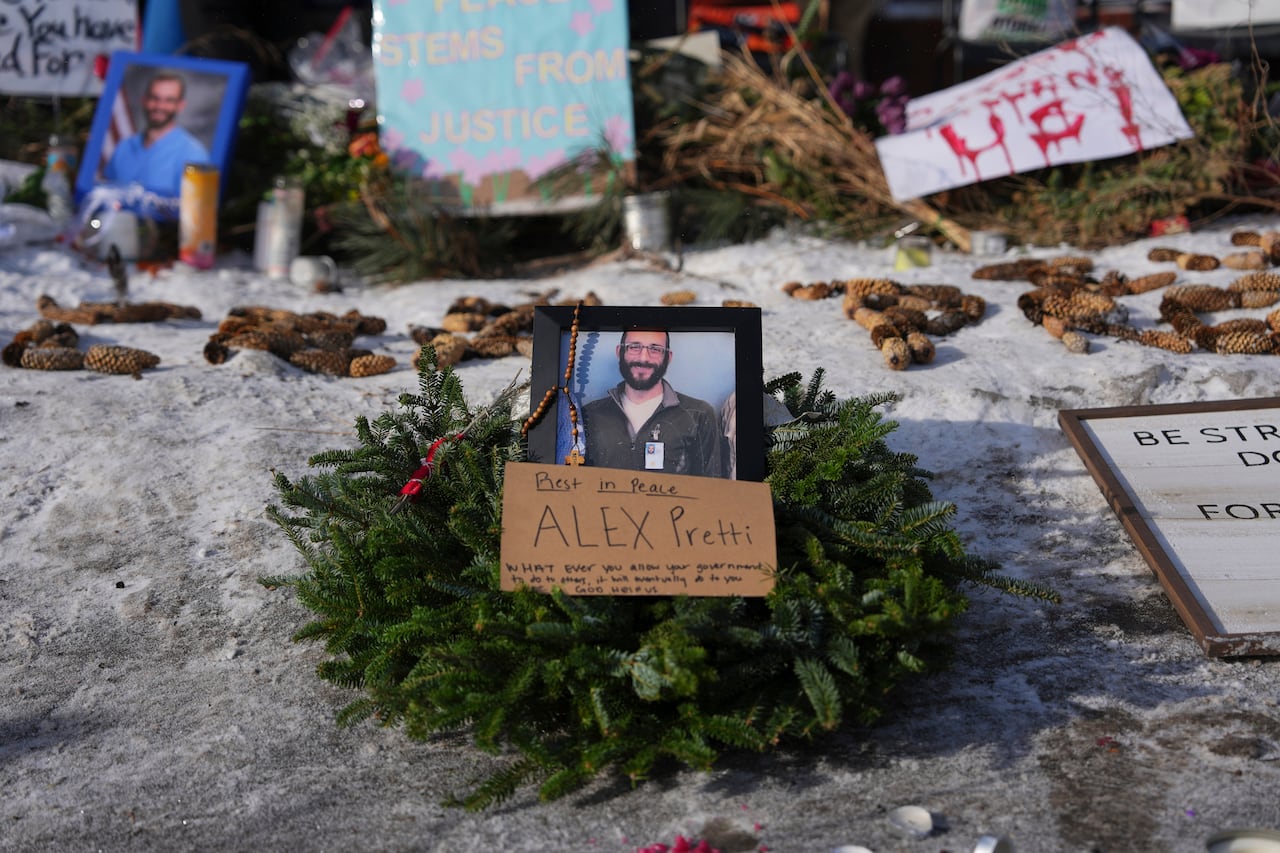 A photo of a man on top of a wreath on a snowy pathway. There is a cardboard sign in front of the pgoto that says 'Rest in Peace ALEX Pretti.'