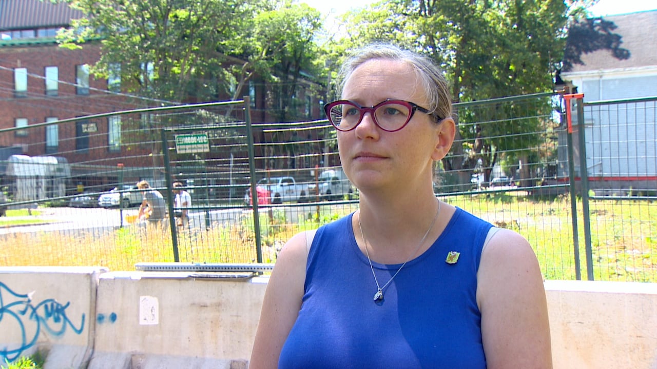 A white woman with blonde hair in a ponytail, red glasses and and blue sleeveless shirt stands in front of an empty lot. There is fencing behind her