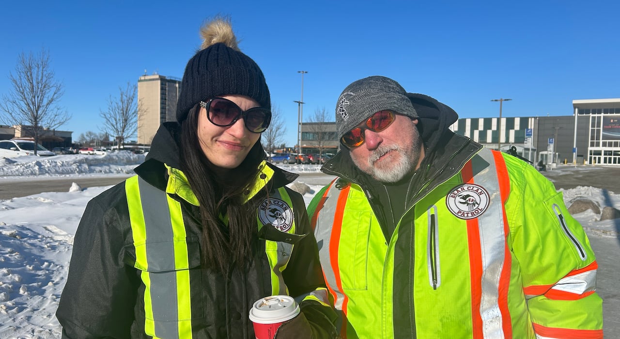 Woman in black and yellow construction jacket stands and black winter hat holds a coffee cup and stands next to a man in a yellow and orange construction jacket on a winter day