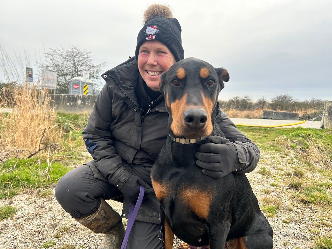 Mia Ribeck is a white woman in a Hello Kitty black touque with a pom pom on top. She's wearing a black parka and thigh high rubber boots. She is squatting next to her dog Greta, a large black Doberman.