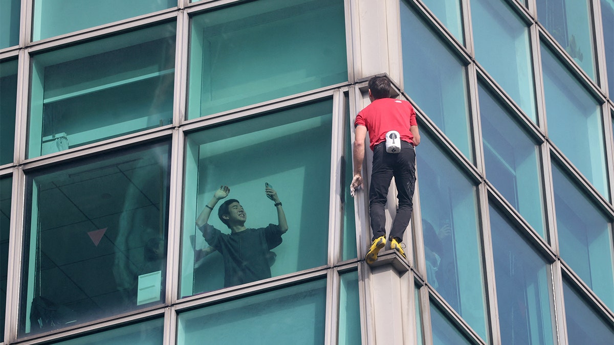 A man films a climber scaling the exterior of a skyscraper from a nearby building.