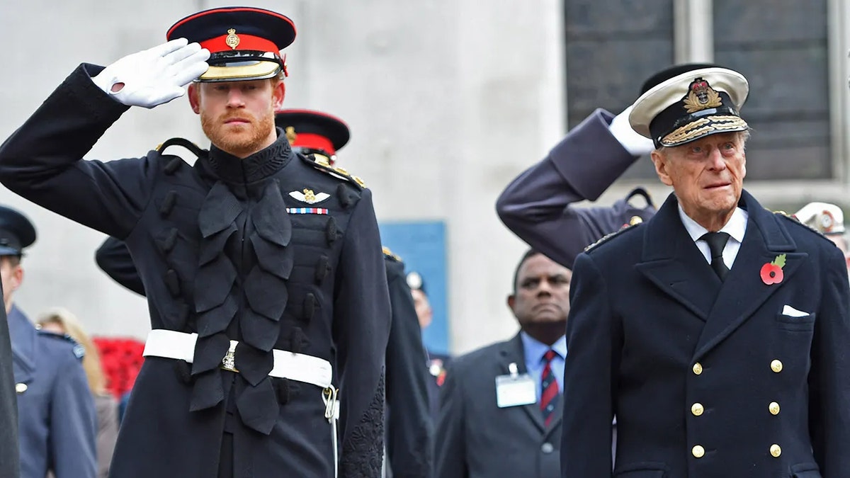 Prince Harry saluting in uniform while Prince Philip, also in uniform, looks on.