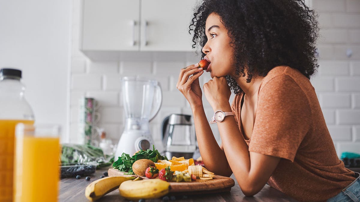Woman eating strawberry in front of plate of fruit in the kitchen.
