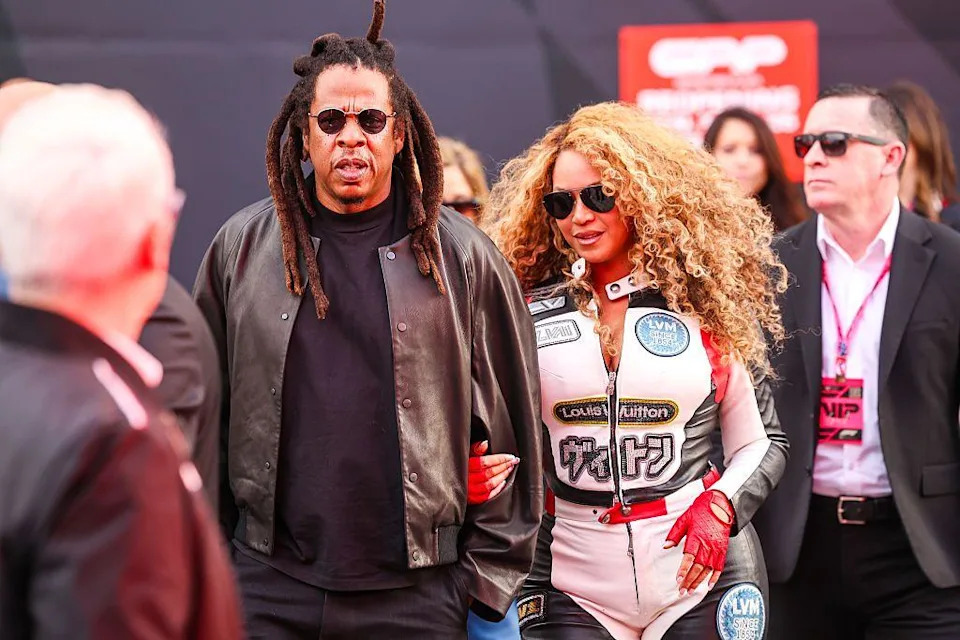 Picture of Jay-Z wearing a brown jacket with a t-shirt underneath and Beyoncé wears a white racing suit with red and black accents.