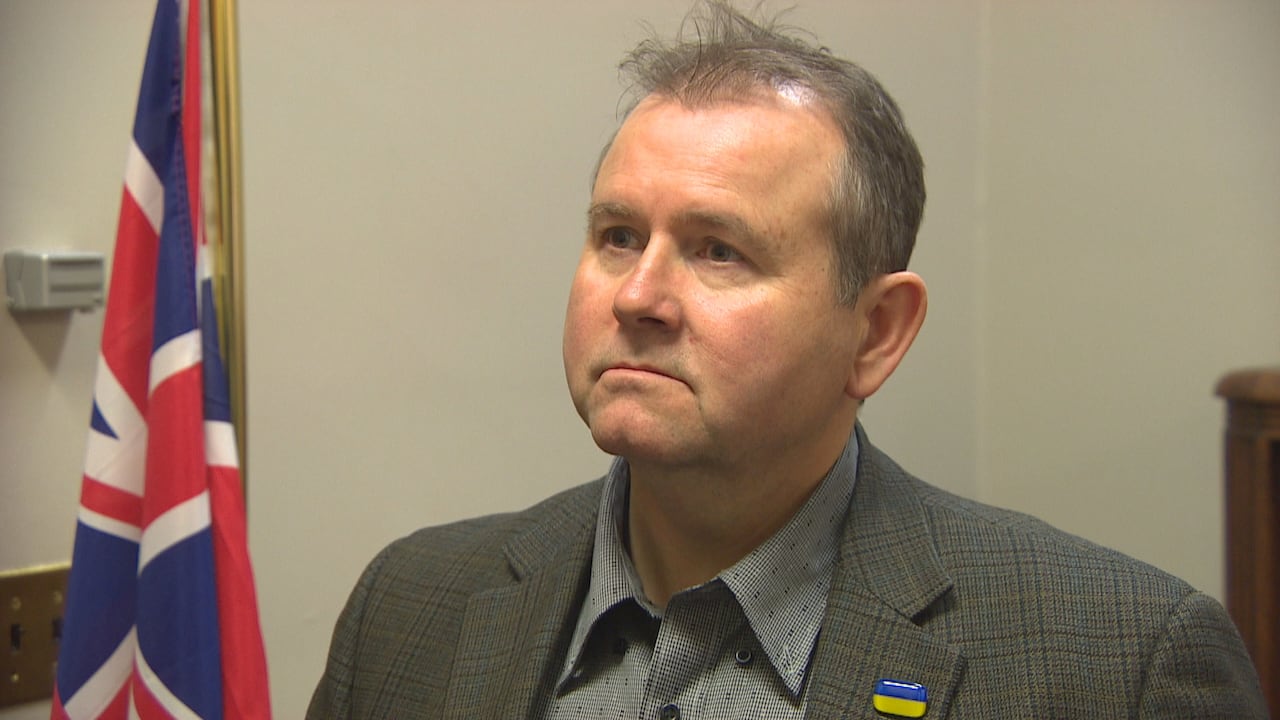 Man with grey hair wears a grey suit with a yellow and blue flag on the lapel