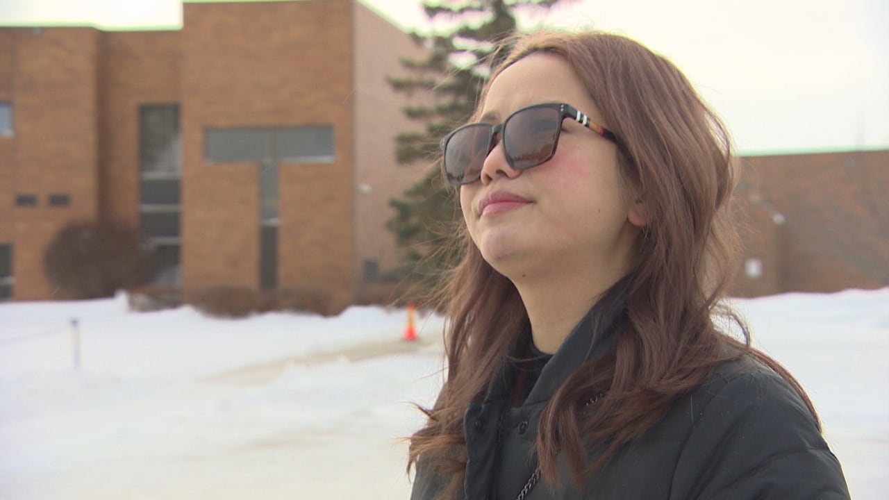 woman with long brown hair wears sunglasses and a black winter coat outside a brick building in the winter