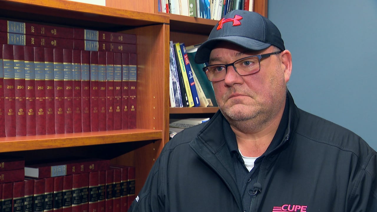 A man with classes wears a black Under Armour ball cap and a black jacket. He is standing in front of a row of library books. 