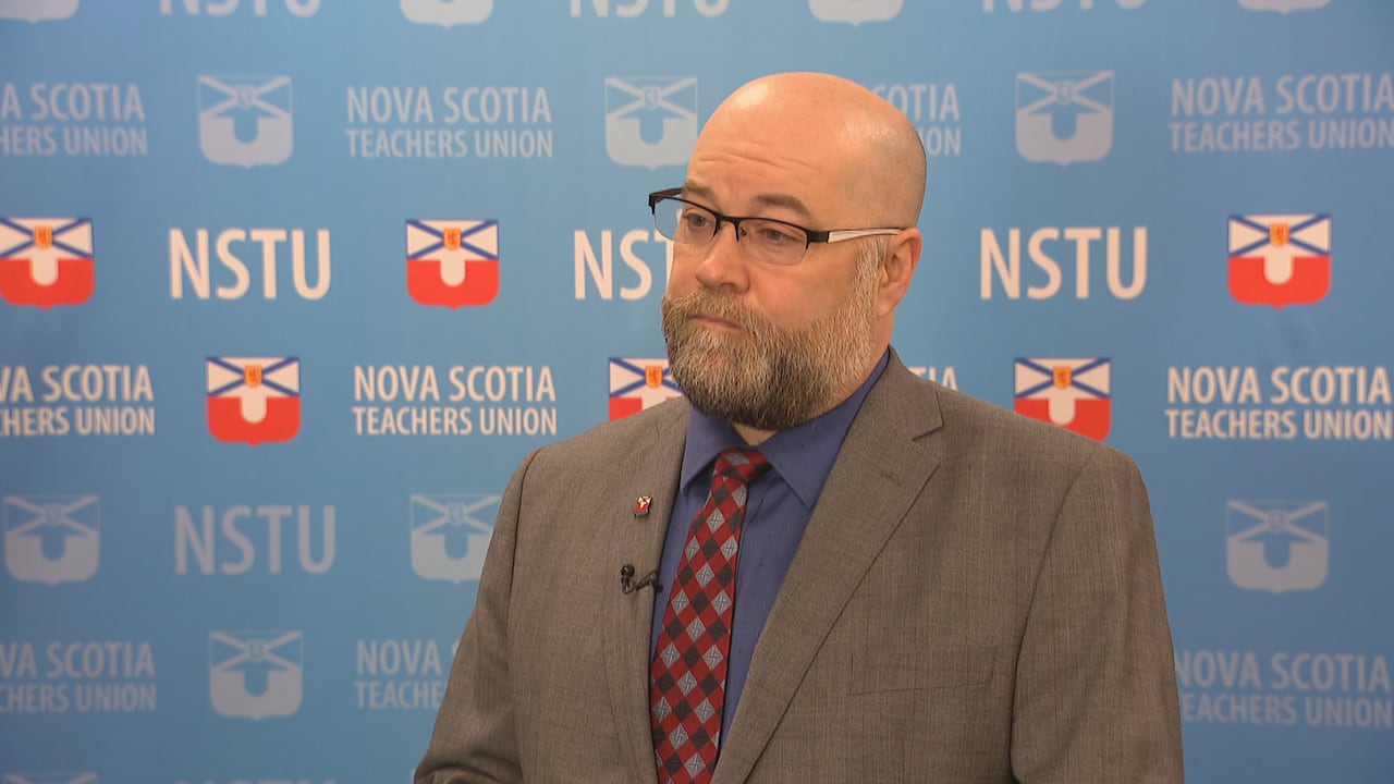 A man wears glasses and a grey suit. He stands in front of a backdrop that displays the Nova Scotia Teachers Union logo. 