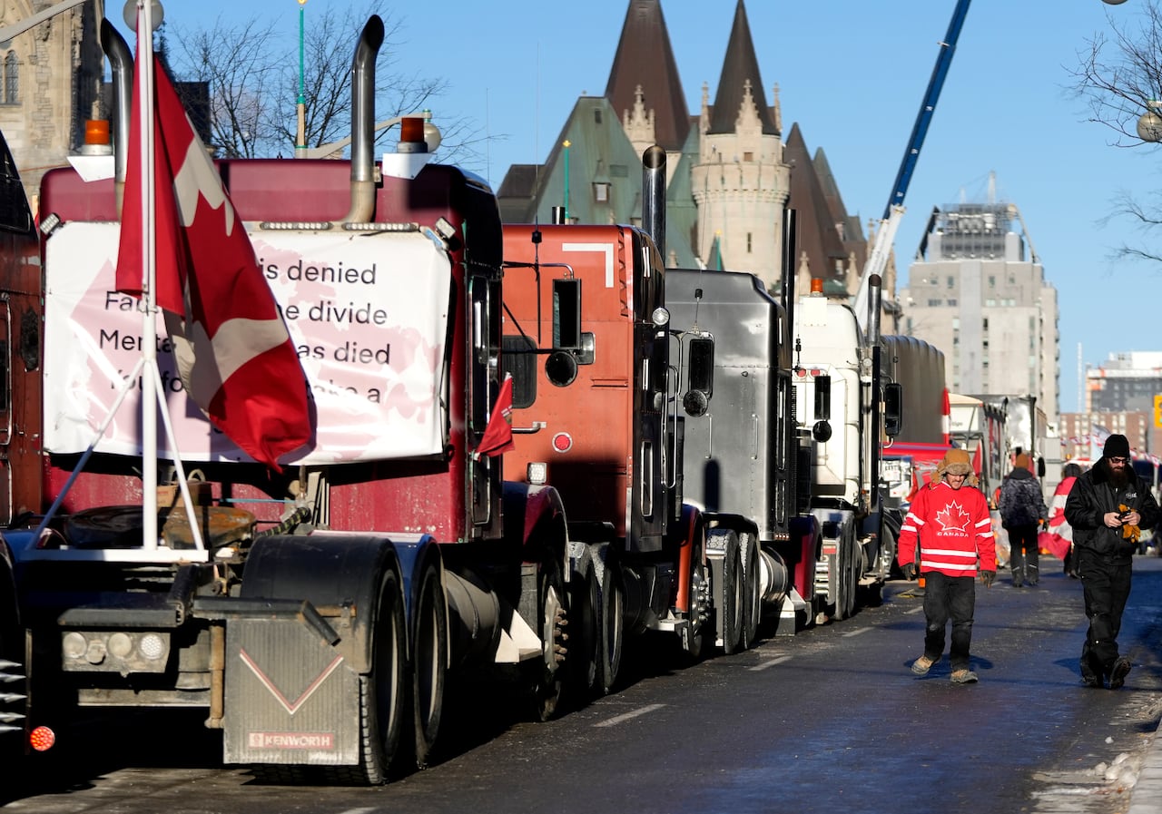 A few men in winter outfits walk beside a row of transport trucks that are lined up in the middle of a road.