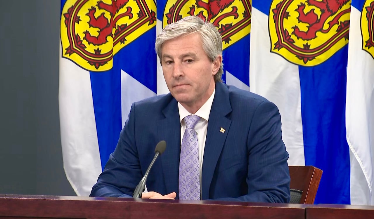 A man in a suit and tie with white hair sits in front of Nova Scotia flags.