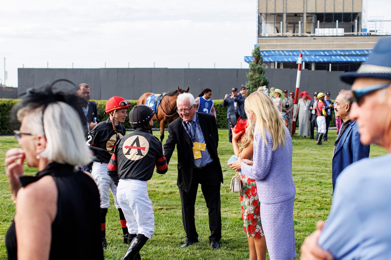 An elderly man shaking a jockey's hand at a racetrack before a horse race, with a racehorse and other people nearby.
