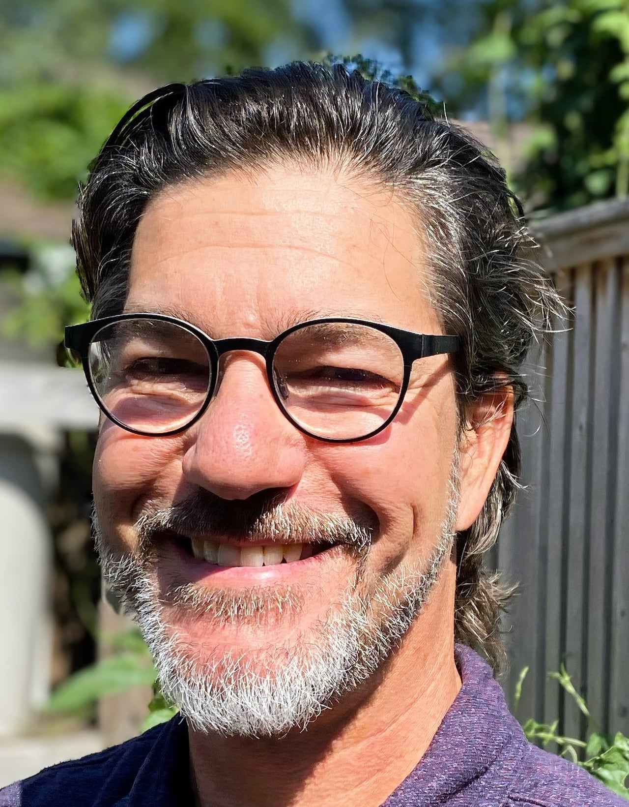 A man with glasses and a short beard smiles outside.