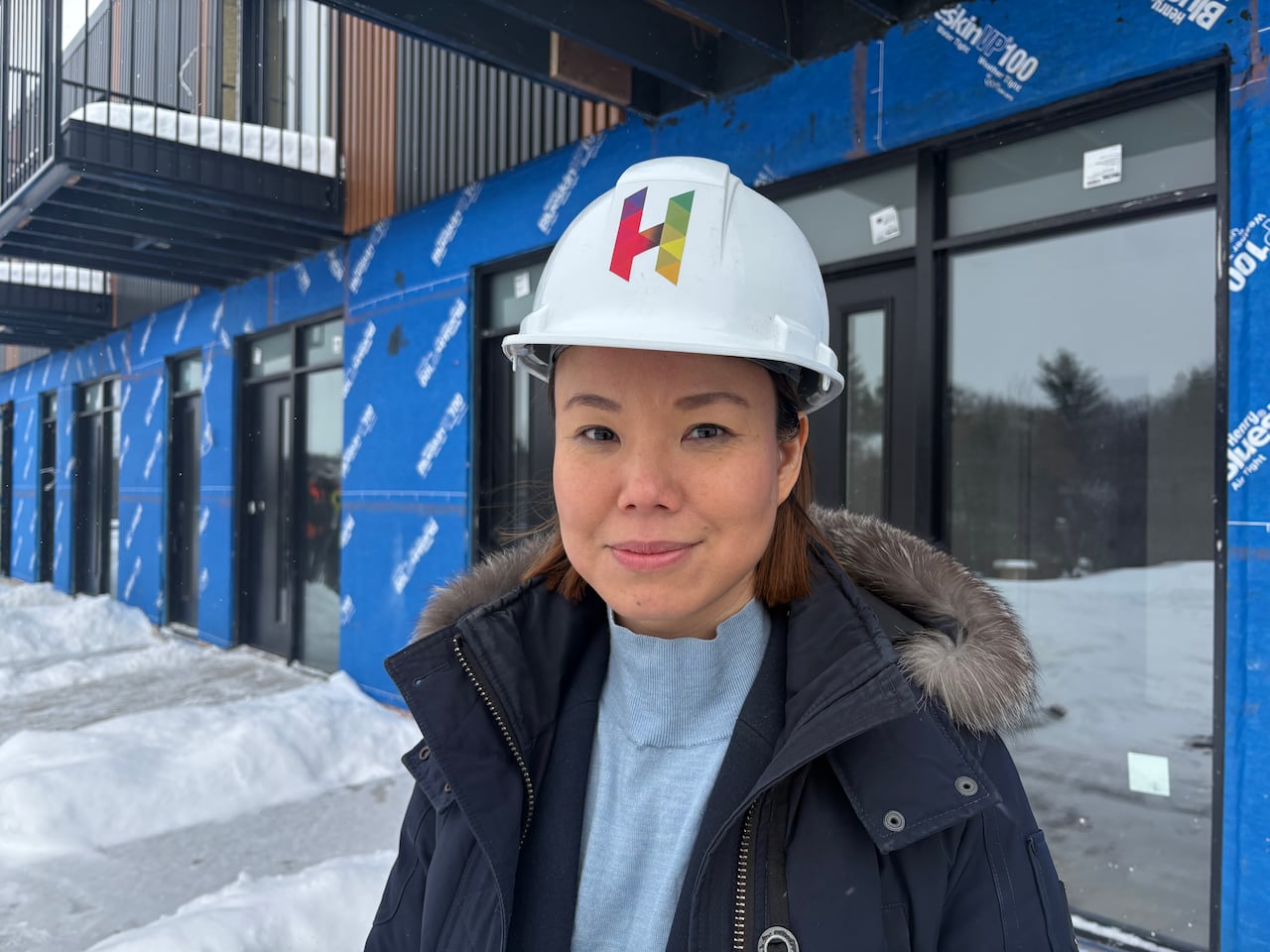 A woman in a parka wearing a white hard hat with a colourful H on it stands in front of a townhome that's under construction.