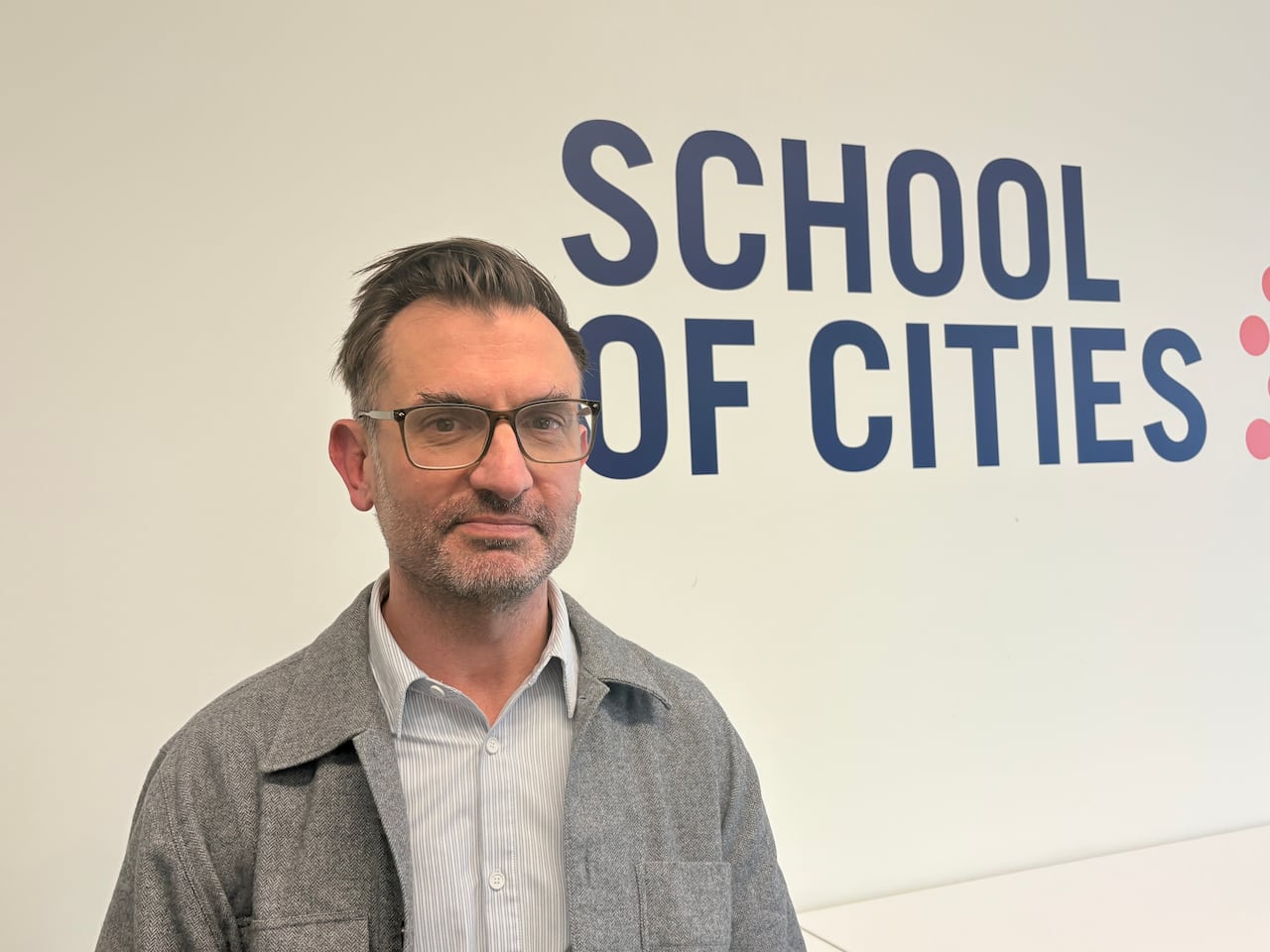 A man with short dark hair and glasses stands in front of a wall featuring a sign that says school of cities.