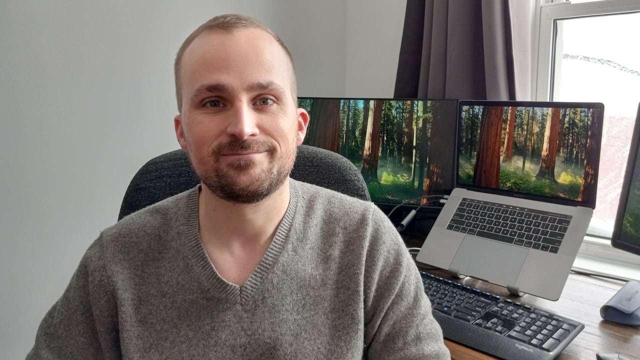Man in grey sweater sitting at a desk with two laptops behind him. 