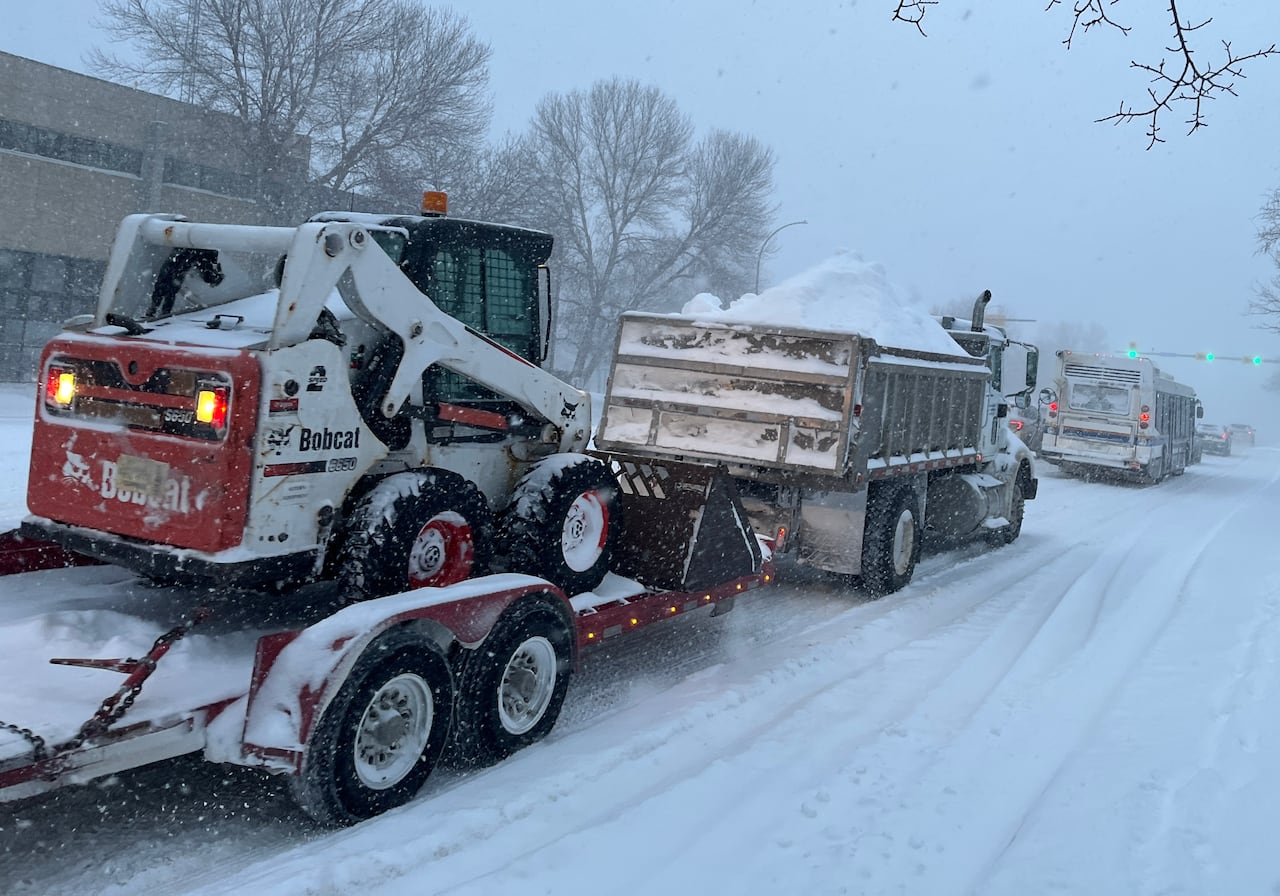 A dump truck piled with snow pulls a trailer carrying a front end loader down a snow street.