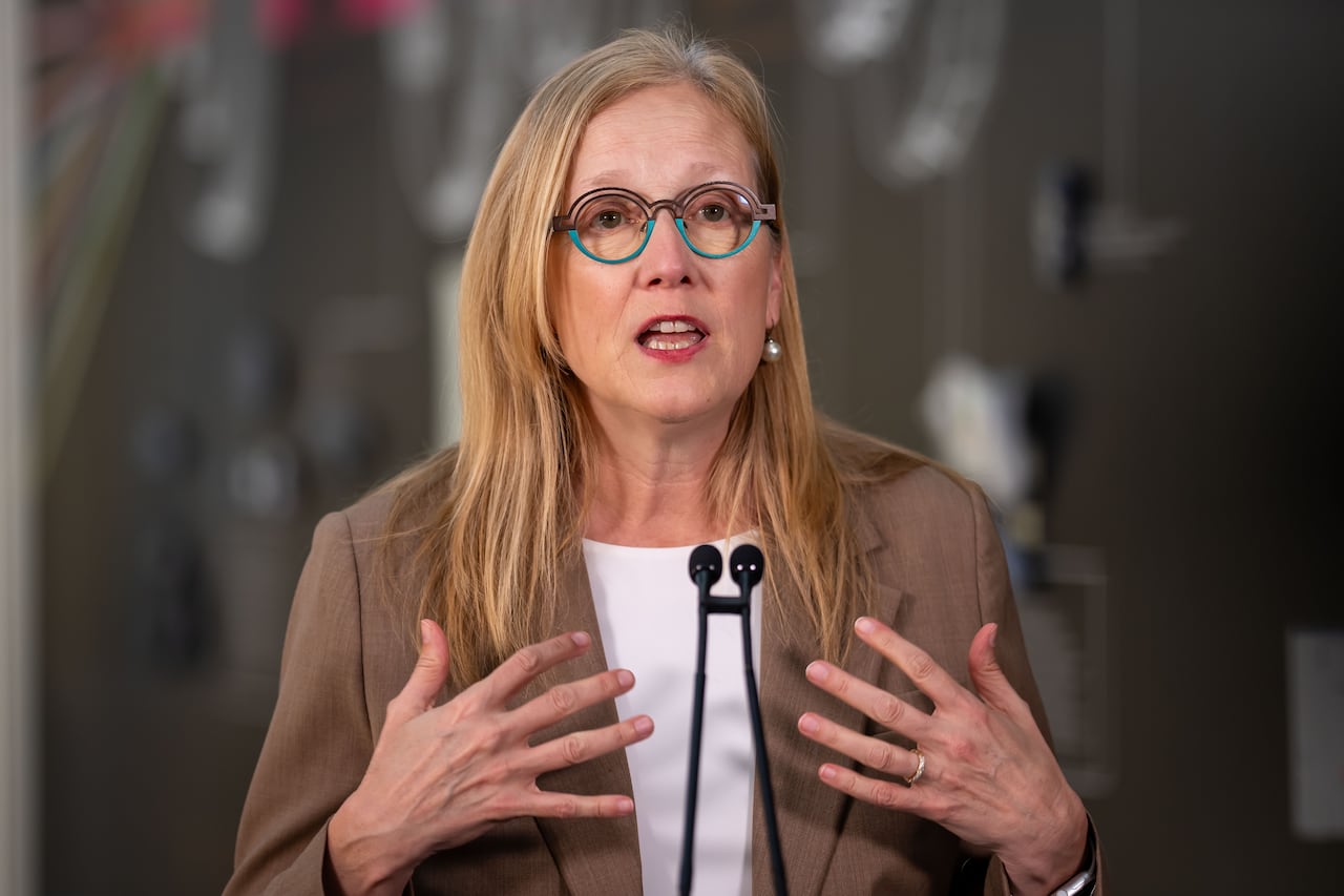 A woman with dirty-blond hair and round eyeglasses speaks at a podium, gesturing with both hands.