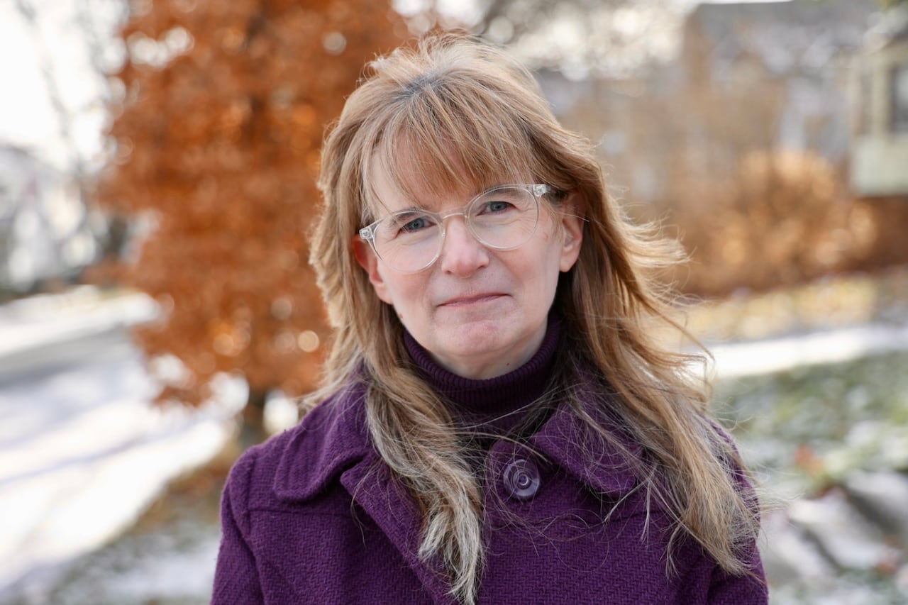 A woman wears glasses and a purple coat. She poses for a photo outdoors in the winter. 