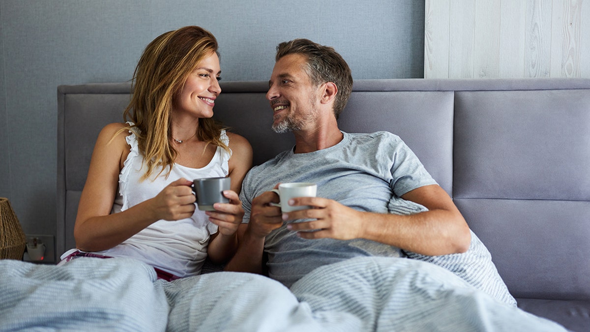couple smiles at eachother in bed holding mugs