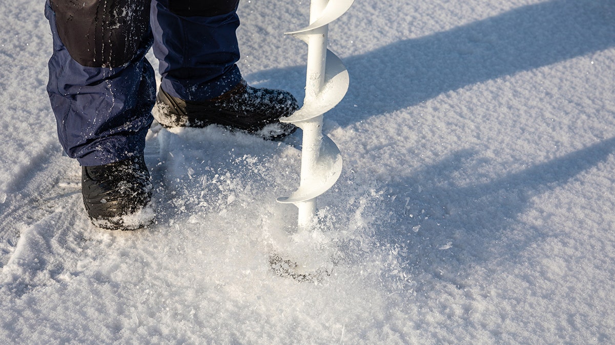 Person drilling a hole in ice with a manual ice auger for ice fishing on a frozen lake.