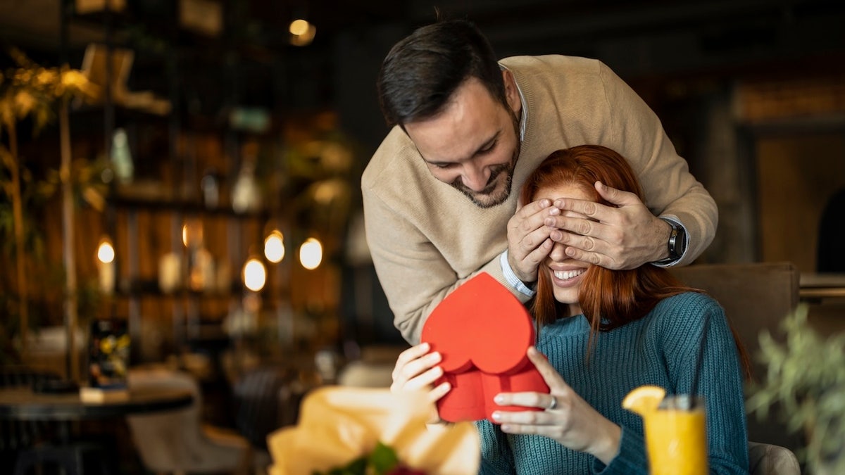 woman receiving valentine's day gift from male partner, who has her eyes covered to keep surprise