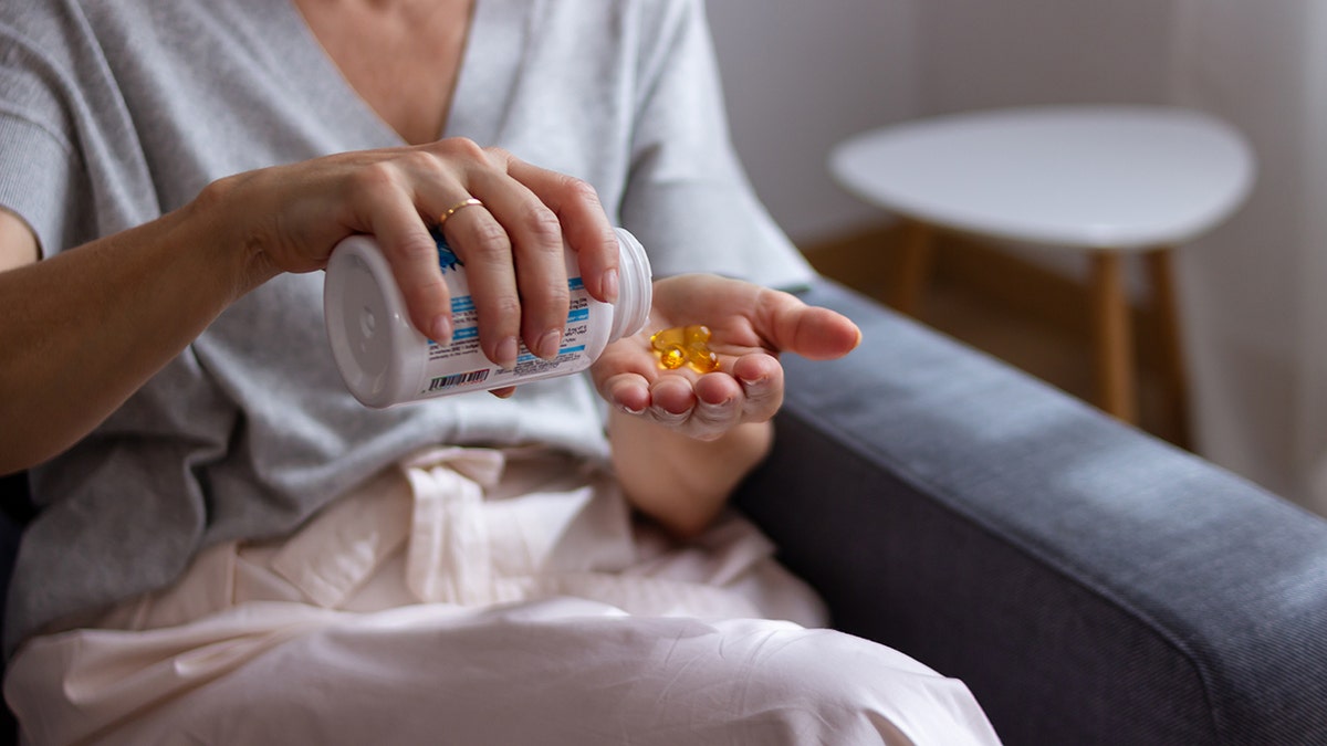 Person pouring yellow softgel supplements from a bottle into their hand while sitting on a couch.