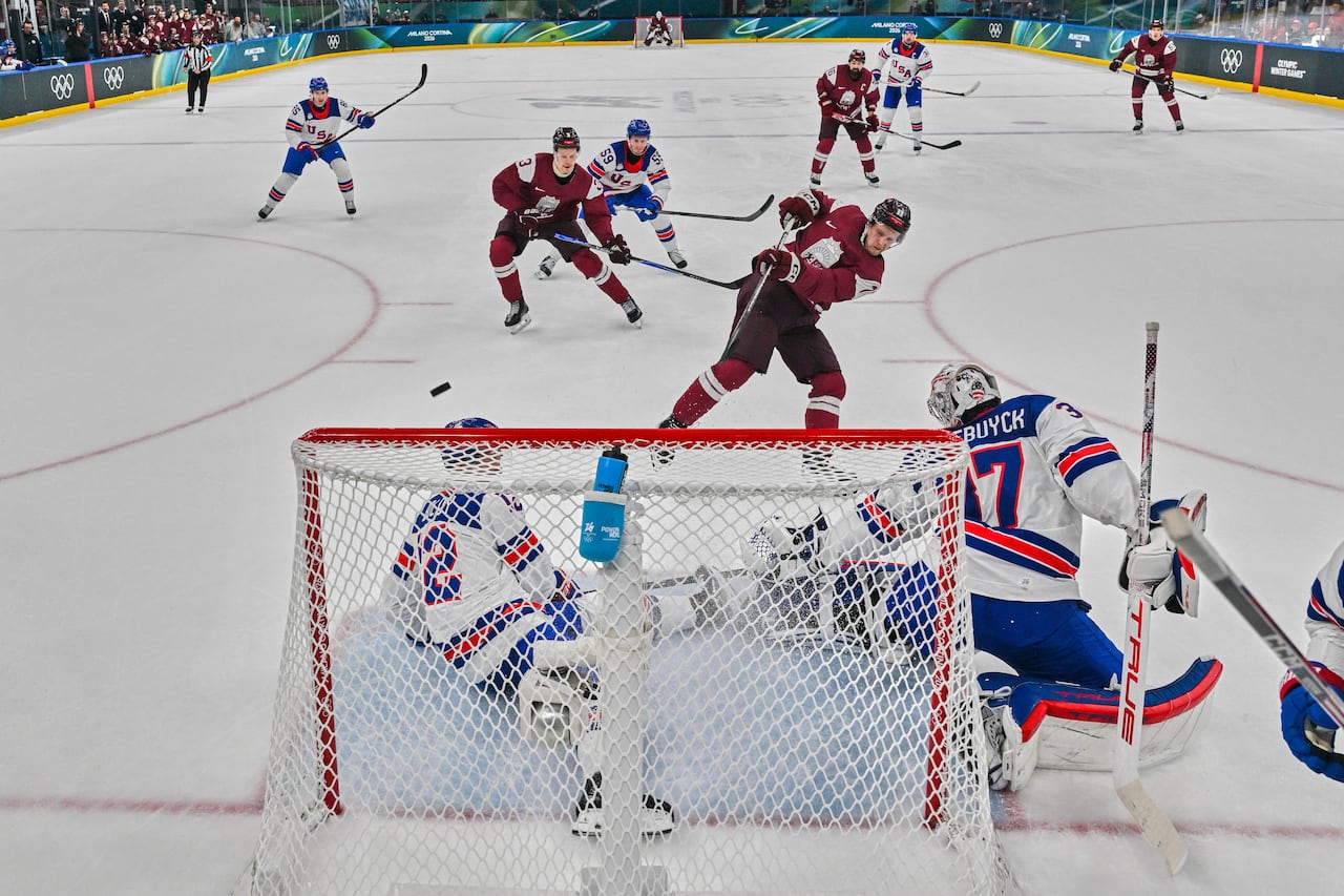 A men's hockey player wearing red shoots at the net of a goalie wearing white.