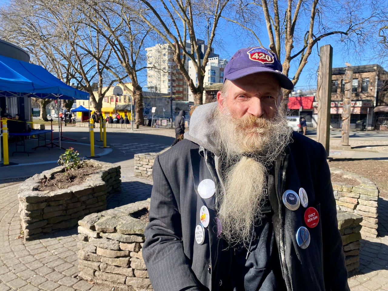 An old man with a long white beard stands in a sunny plaza, wearing a purple cap and a jacket covered in buttons, including one that reads “Evict Ken Sim.”
