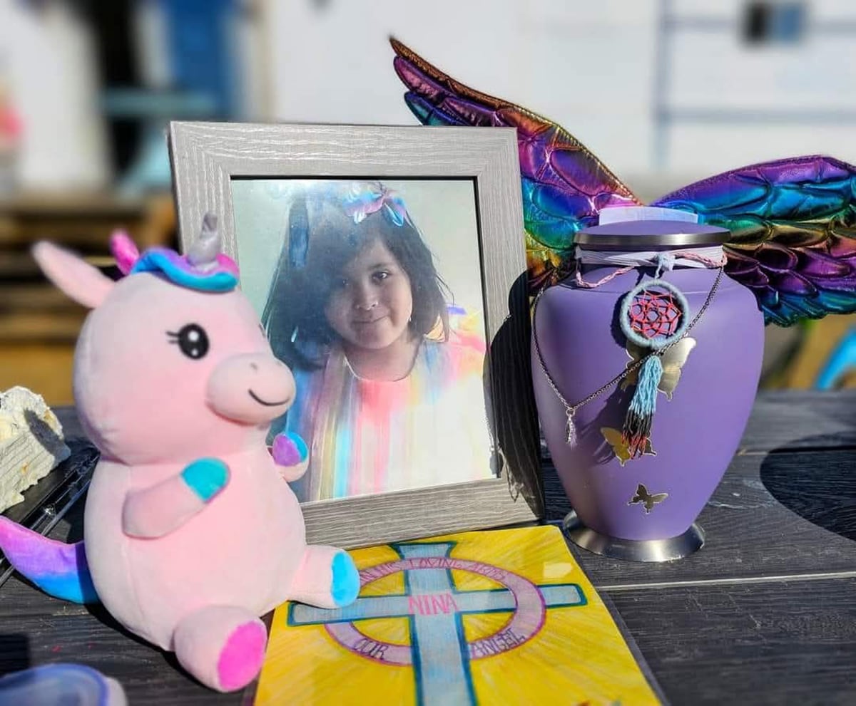 A framed photograph of a little girl with dark hair and striped shirt is surrounded by stuffed animals and an urn.