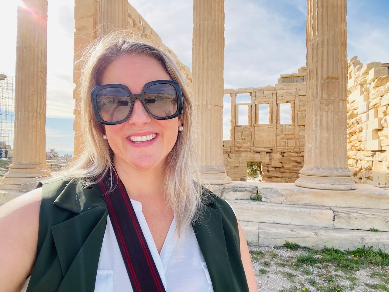 Woman with blonde hair and sunglasses. Behind her is a ruin.