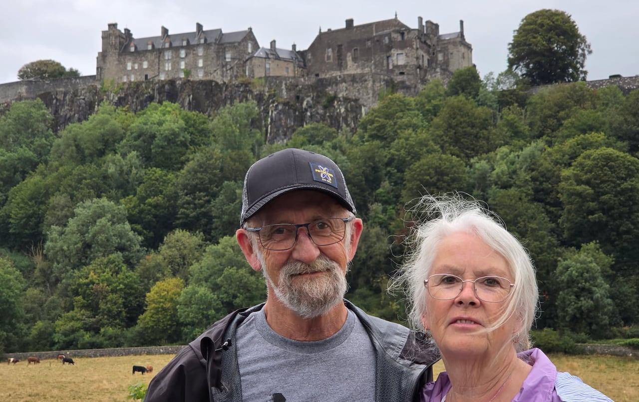 man and a woman, both with white hair, stand below a stone castle on a hill