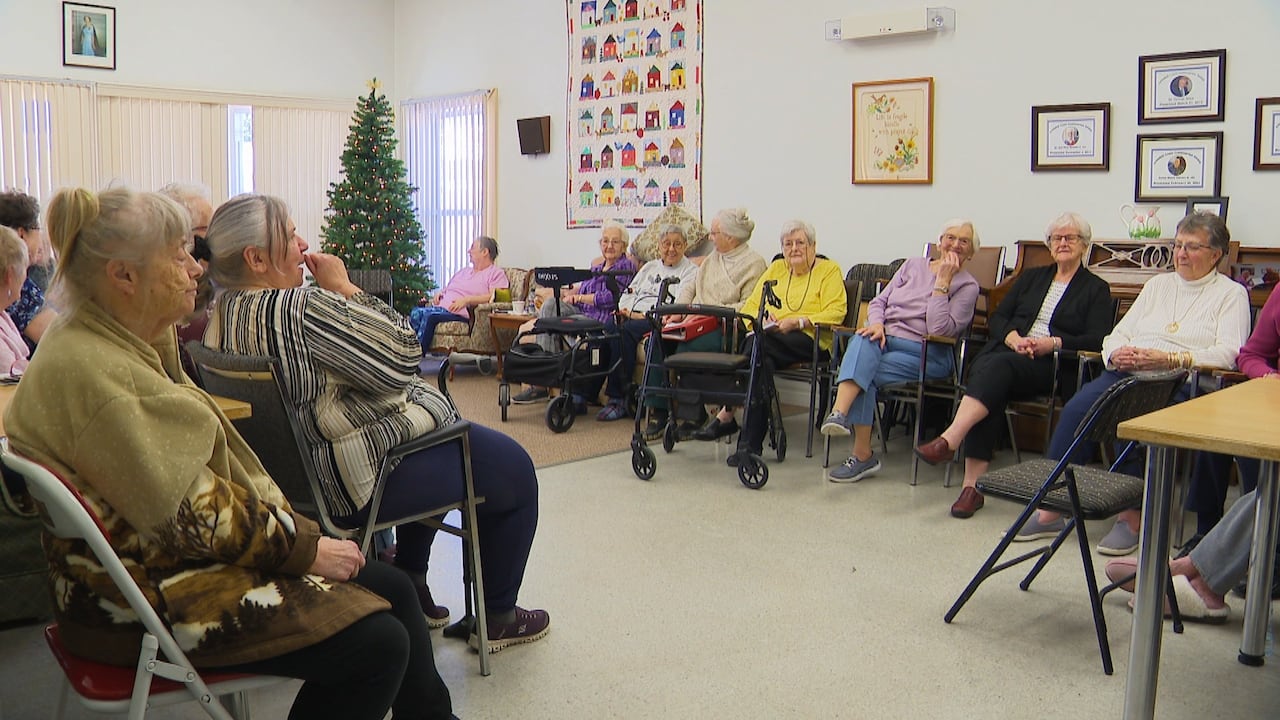 A group of seniors sit in a semi circle around the common room of their independent living facility. 