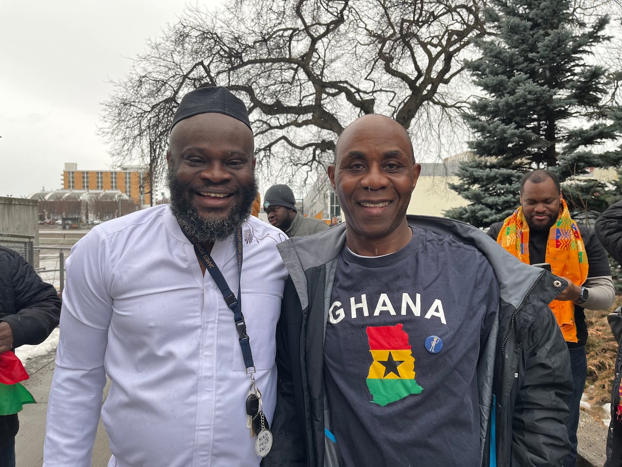 Two men pose together and smile on a rainy day. One is wearing a shirt with the Ghana flag. 
