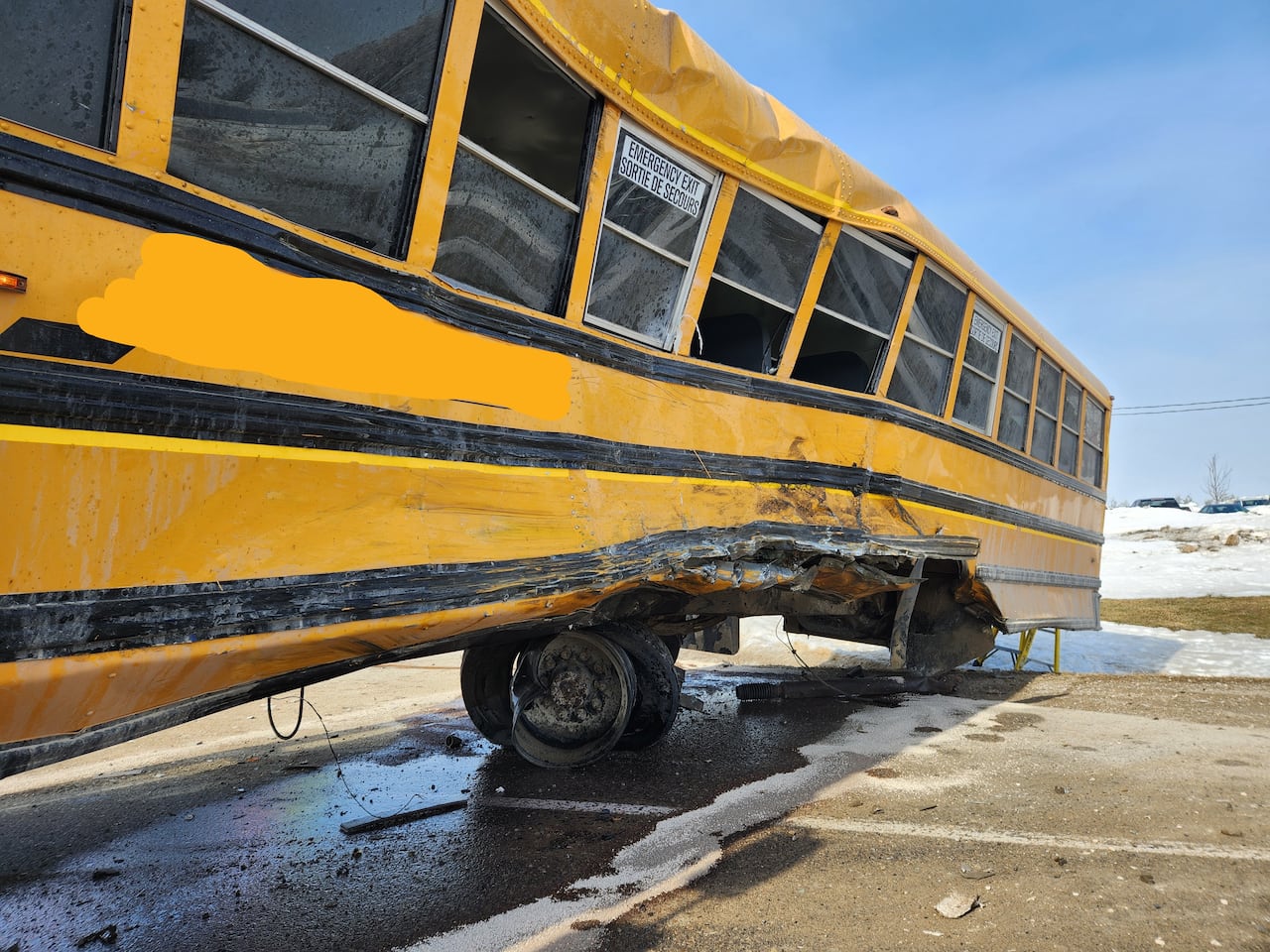 Close-up of the crushed driver’s side of a yellow school bus, with metal bent inward above the rear wheel after a collision.