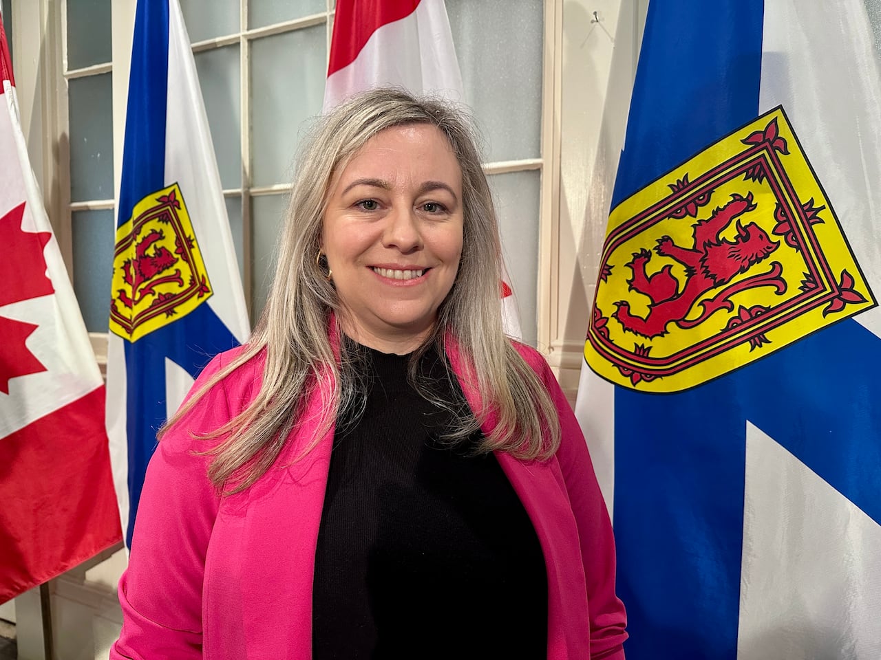 A woman with blonde hair is wearing a black shirt under a bright pink blazer stands in front of a four flags.