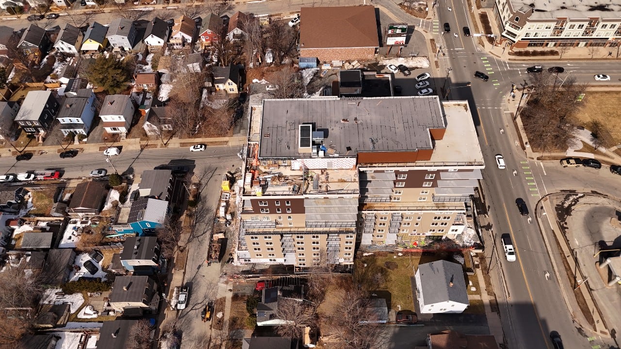 The roof and penthouse of a tall apartment building under construction is seen from above, with surrounding single-family homes and streets.