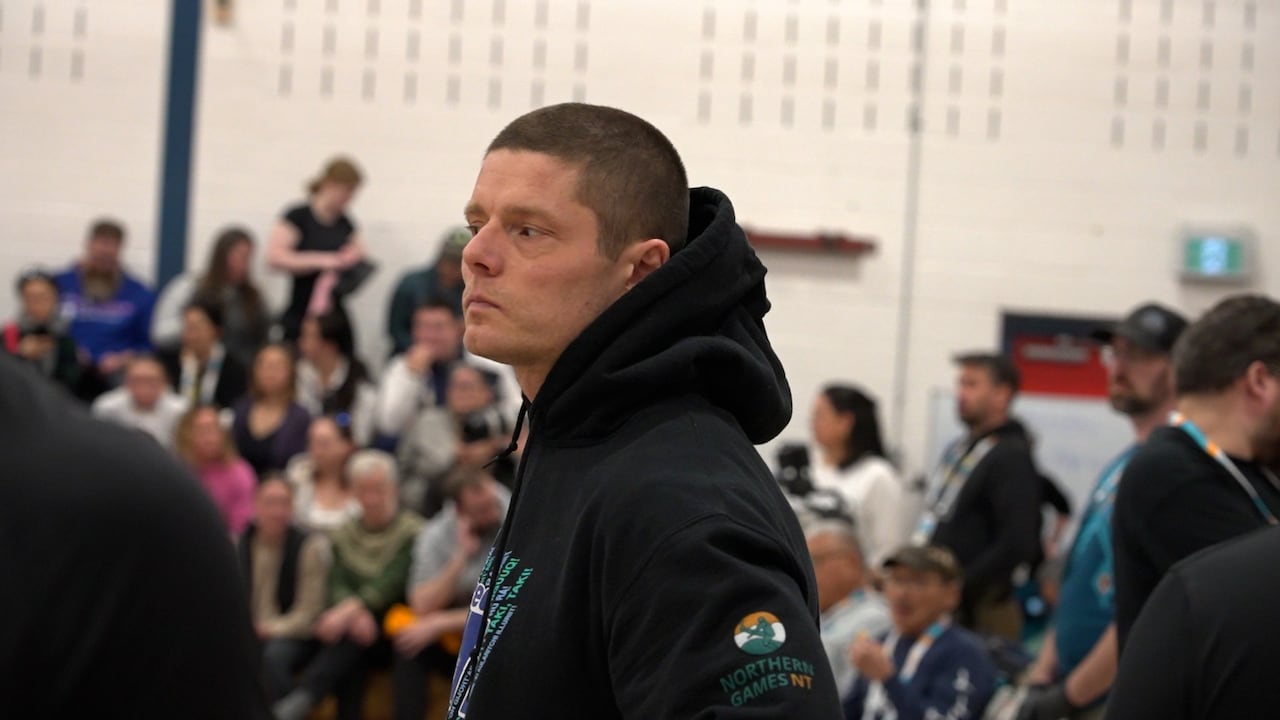 A man in a black hoodie stands and looks away from the camera in a room full of people.