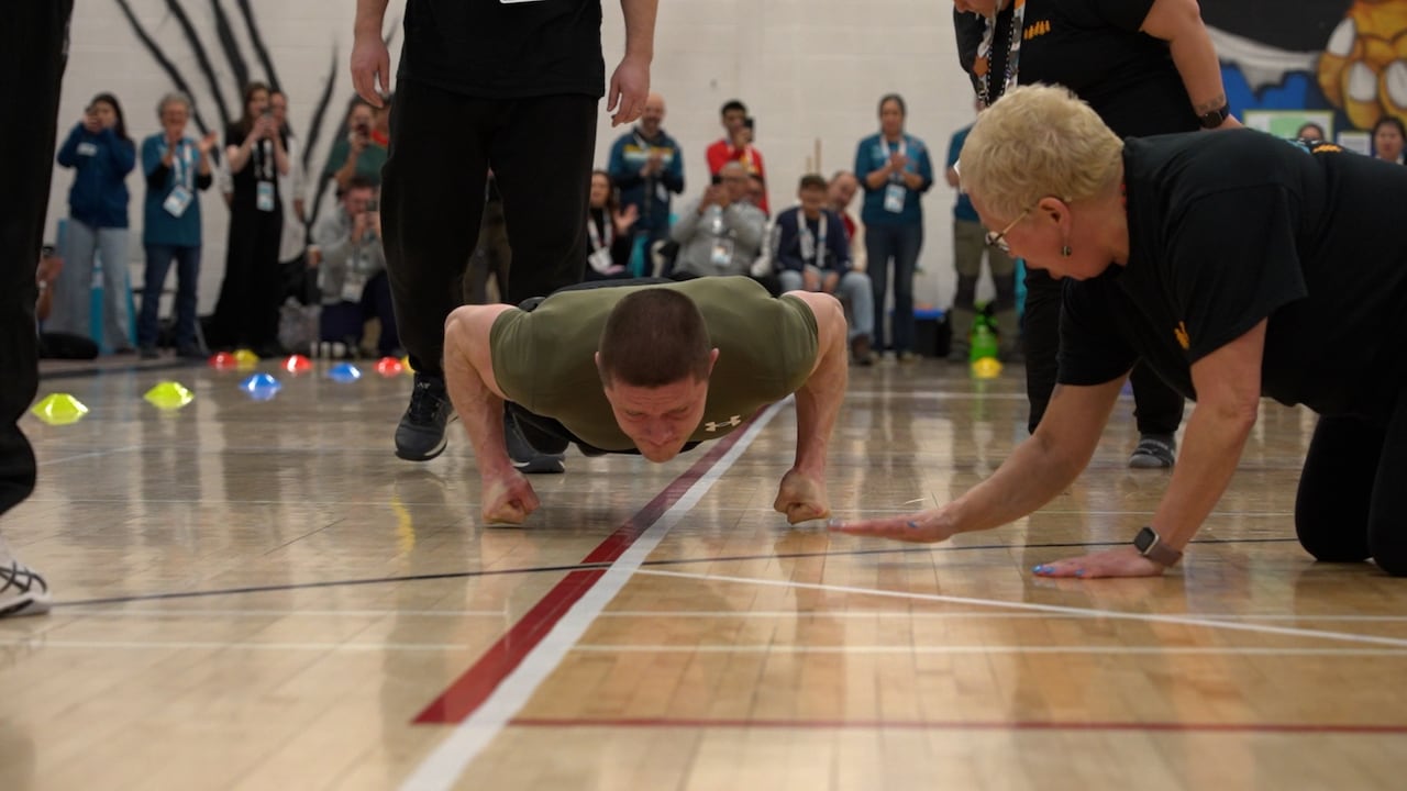 A man hovers a few inches off the ground in a plan position with his fists clenched as a women pounds the gym floor in front of him.