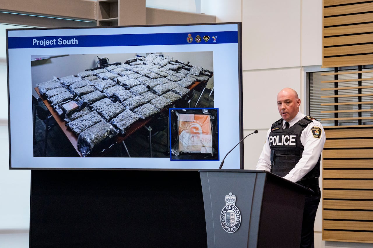 A police chief stands on stage delivering a news conference, next to a digital display.