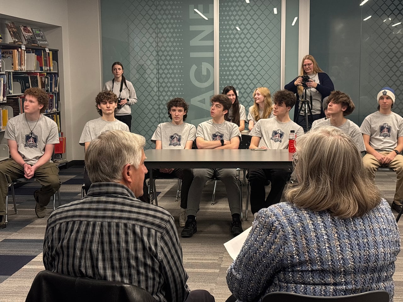 Students wearing Artemis II Explorers Club shirts sit at a table in a school library while Nancy and Gary Hansen speak with them during a visit to Ingersoll District Collegiate Institute.