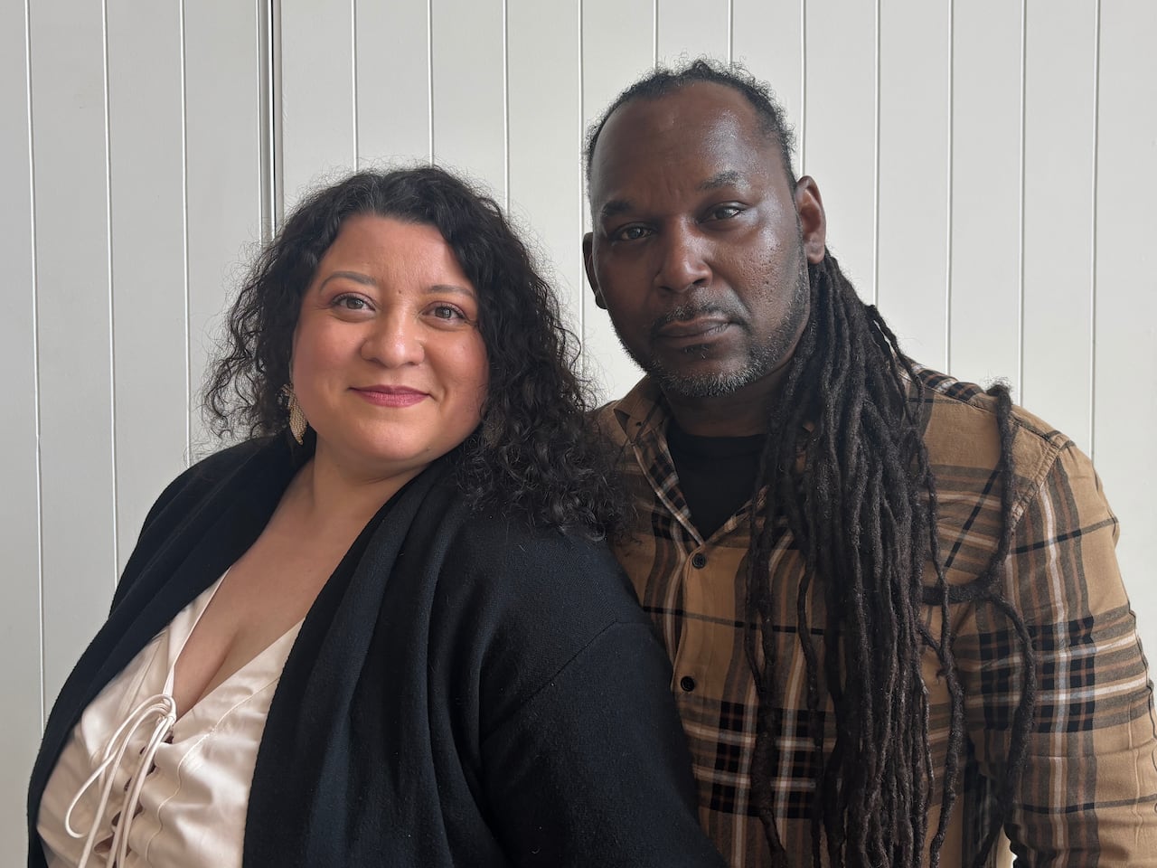 Brenda Guzman and Vencotte “V” Merchant standing side by side facing the camera against a white paneled wall. Guzman wears a cream blouse and black jacket, while Merchant wears a brown plaid shirt and has long dreadlocks over one shoulder.