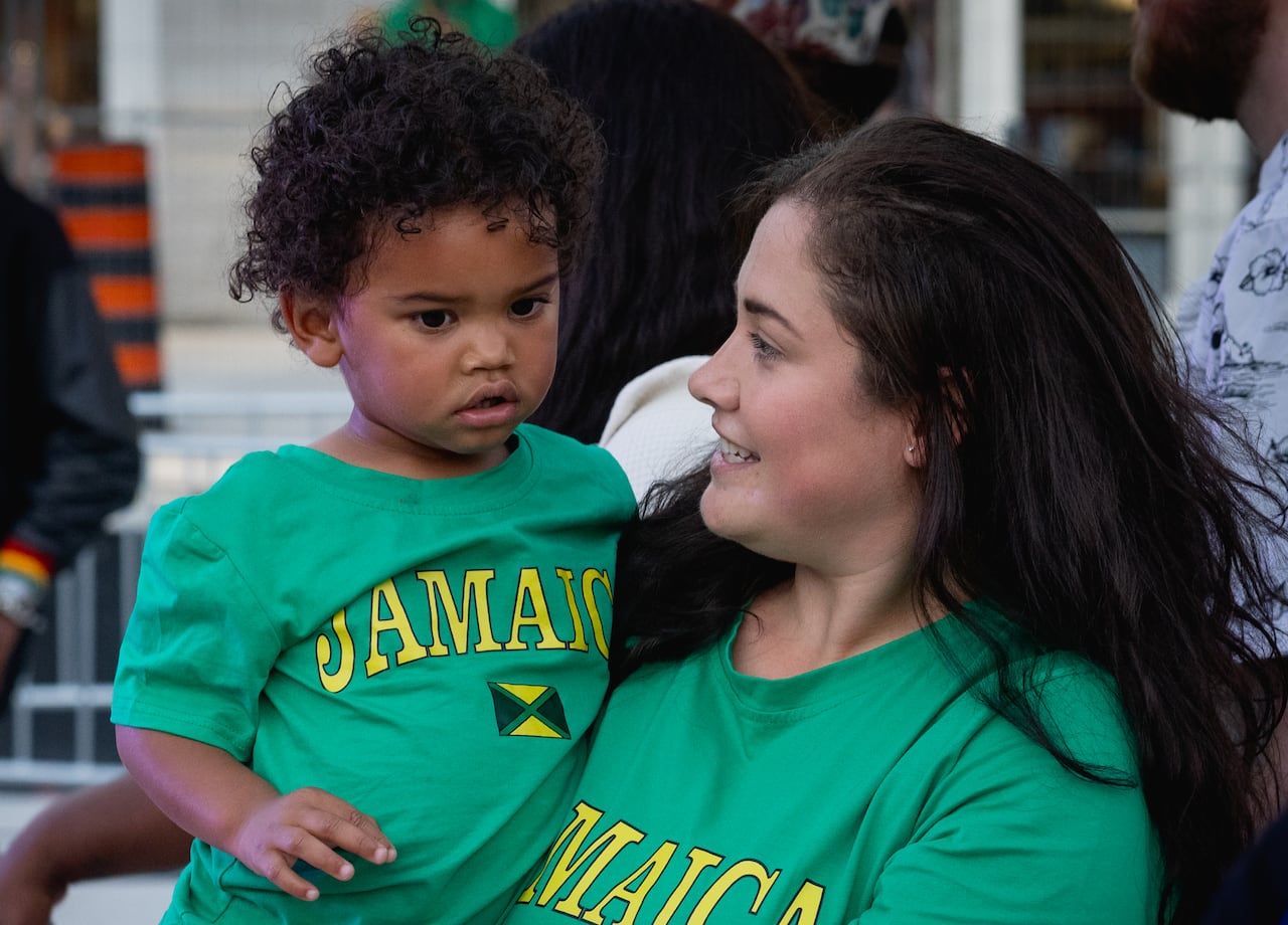 A woman wearing a green Jamaica T-shirt holds a young child in a matching shirt while attending London Reggae Fest, with other festivalgoers in the background.