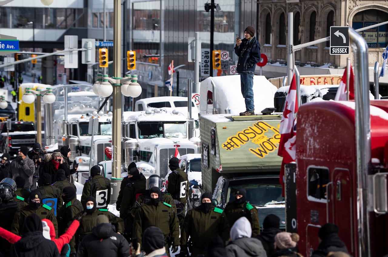 A man stands on semi-truck, one of several parked on a street. Police officers are seen on the ground near the trucks.