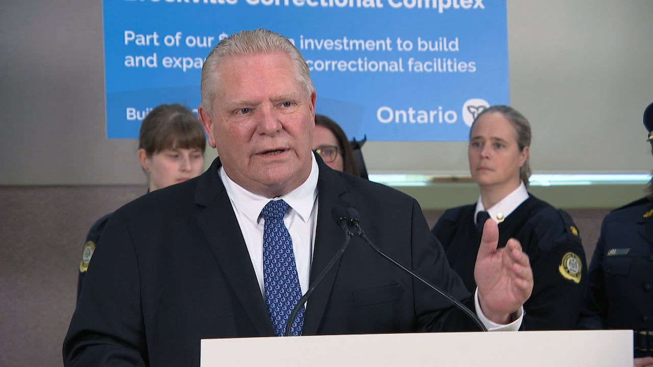 A man with short blonde hair, wearing a suit and blue tie stands in front of a podium. Behind him are jail guards in uniform.