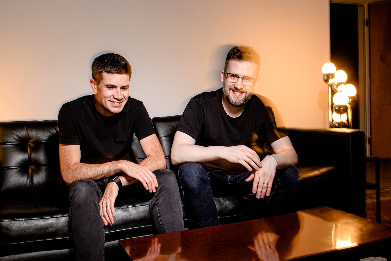 Two smiling people sit on a leather-type fabric couch, illuminated by a lamp to the side. 