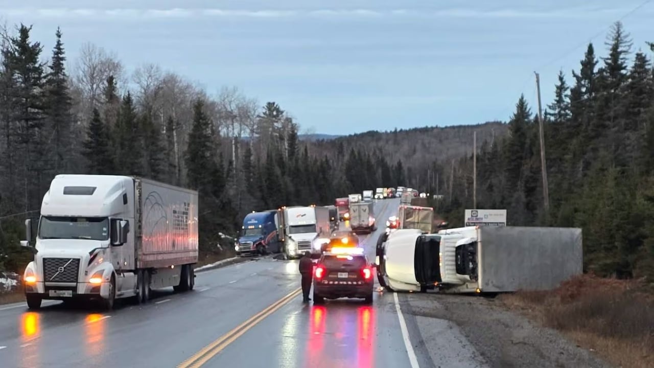 Multiple trucks are seen waiting behind a toppled over transport on a rainy highway.