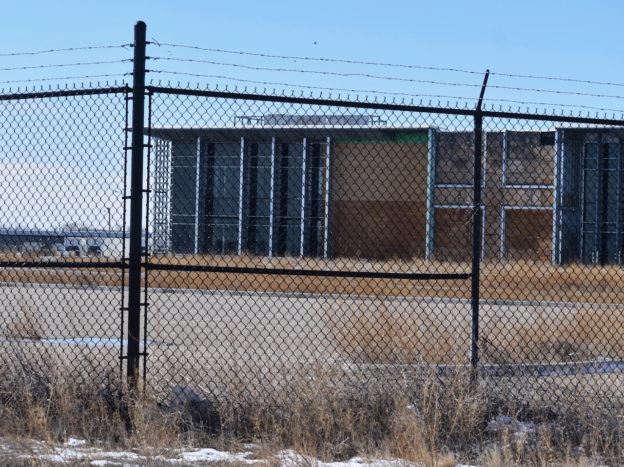 A black chain link fence surrounds a large warehouse that has bare plywood and weeds in an empty parking lot.