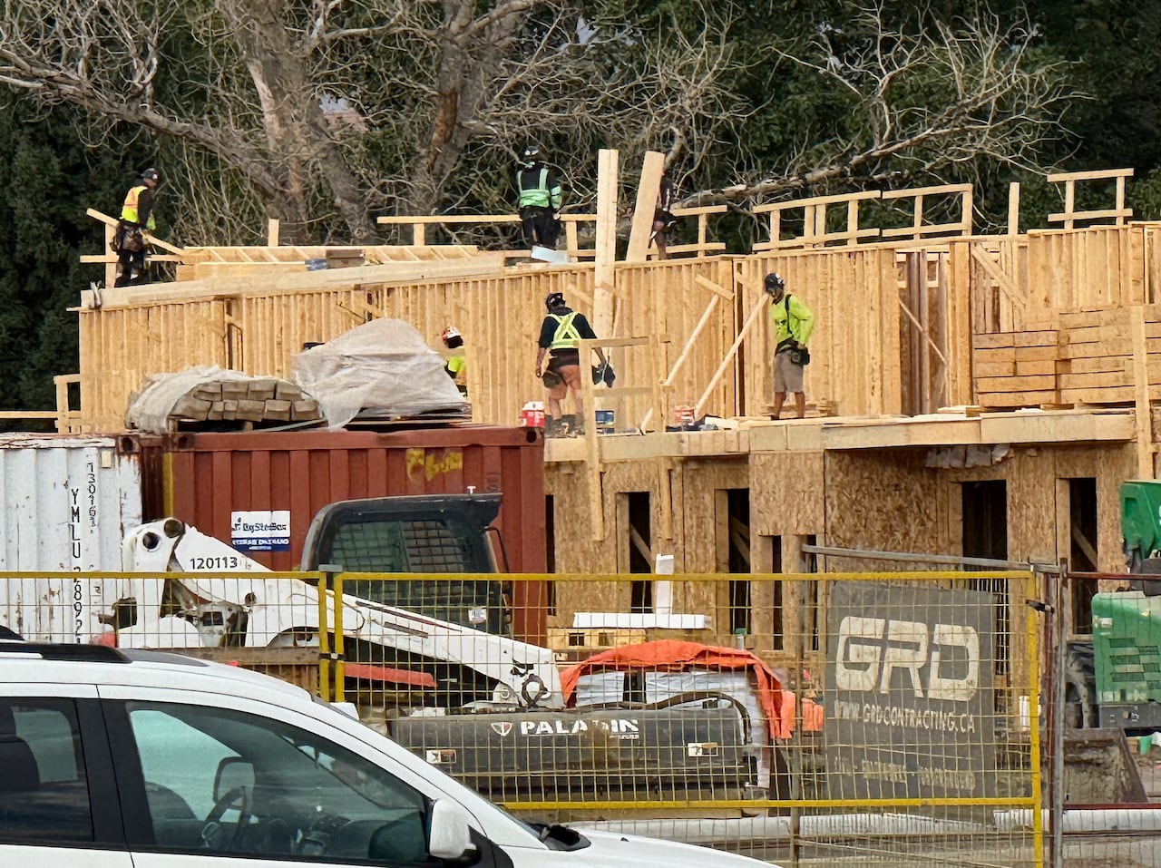 Workers in safety vests walk on a plywood structure behind construction fencing and machinery.