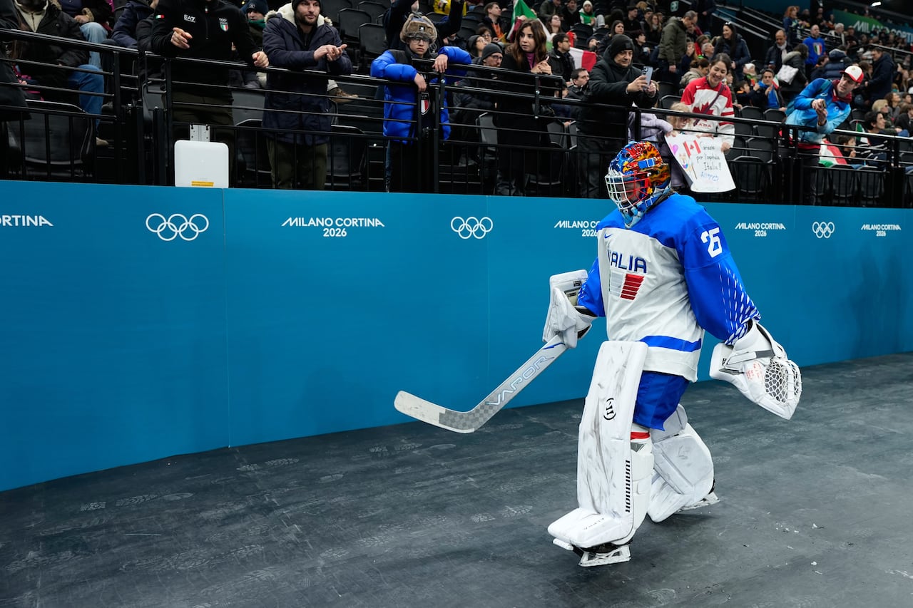 A goaltender walks on to the ice, past a logo showing the Olympic rings.