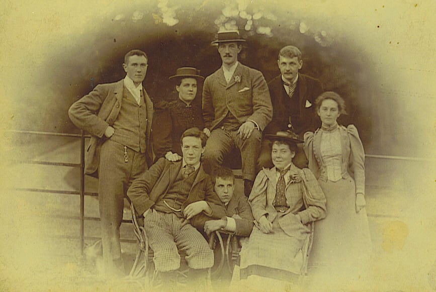 A yellowed old photograph of a well-dressed family huddled together, the edges of the photograph are faded.